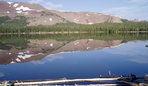 Photo of a lake surrounded by pine trees with a rocky mountain in the background.