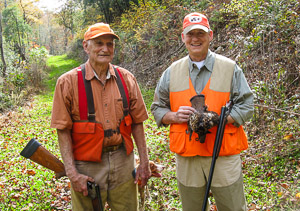 Two smiling old hunters wearing orange vests and hats with shotguns, their dog, and a ruffed grouse.