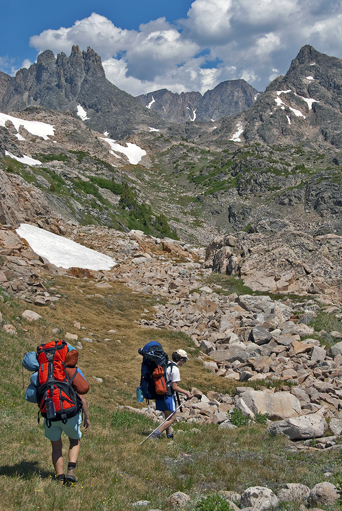 Two people hiking with backpacks on their backs, mountains in the background