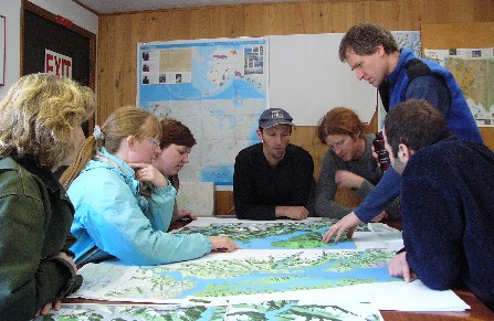 A group of women and men discuss their expedition over maps spread out on a table.