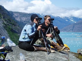 A couple sits on a rock looking through binoculars mounted on tripods.