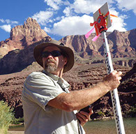 Image of Dr. David M. Merritt holding a survey target, with a body of water and tall desert peaks and mesa's in the background.