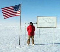 Image of Joel Murray standing between the flag of the United States of America and a location sign.