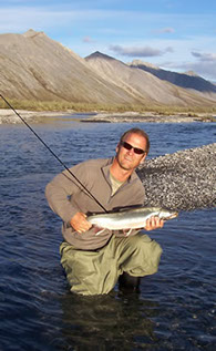 Picture of Brett B. Ropert, PhD National Aquatic Ecologist, standing in a river holding a large fish and a fishing pole. Mountains in background