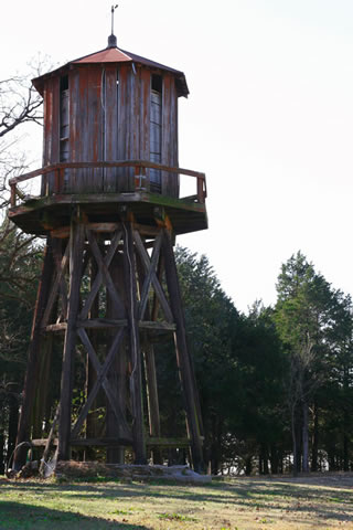 Wooden water tank, with faded red paint, set at the edge of a row of trees.