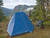 A pitched tent in the foreground with a backpack next to it on the ground with a hiker, facing away from the camera, sitting down in the middle distance enjoying the view of mountains in the background.