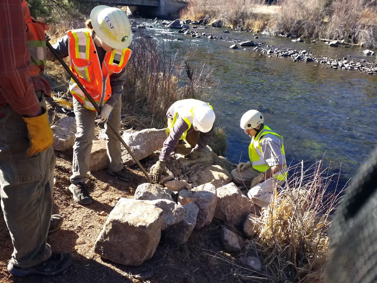 A team of people wearing hard hats, high visibility jackets, and gloves work to move stone blocks on the bank of a river.