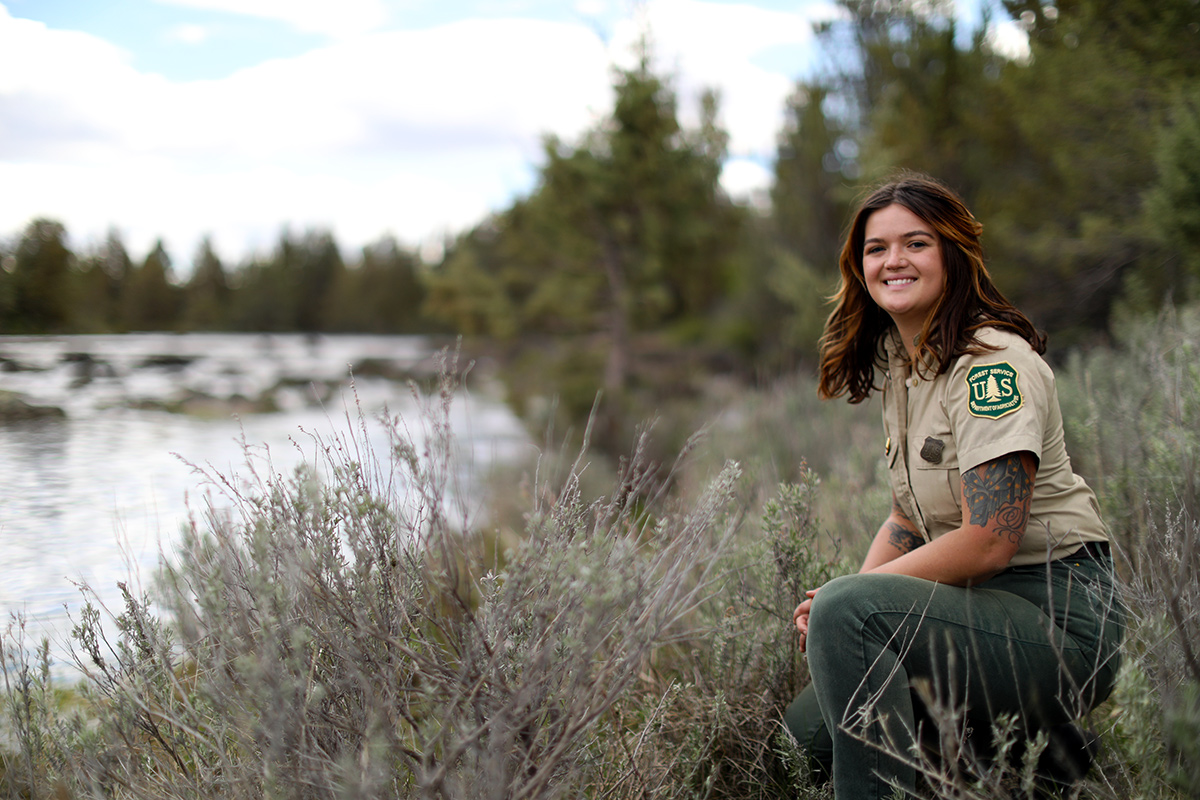 Image of hydrology technician, Kaci Spooner, wearing a USDA Forest Service uniform, kneeling in the short brush and grass next to a river.