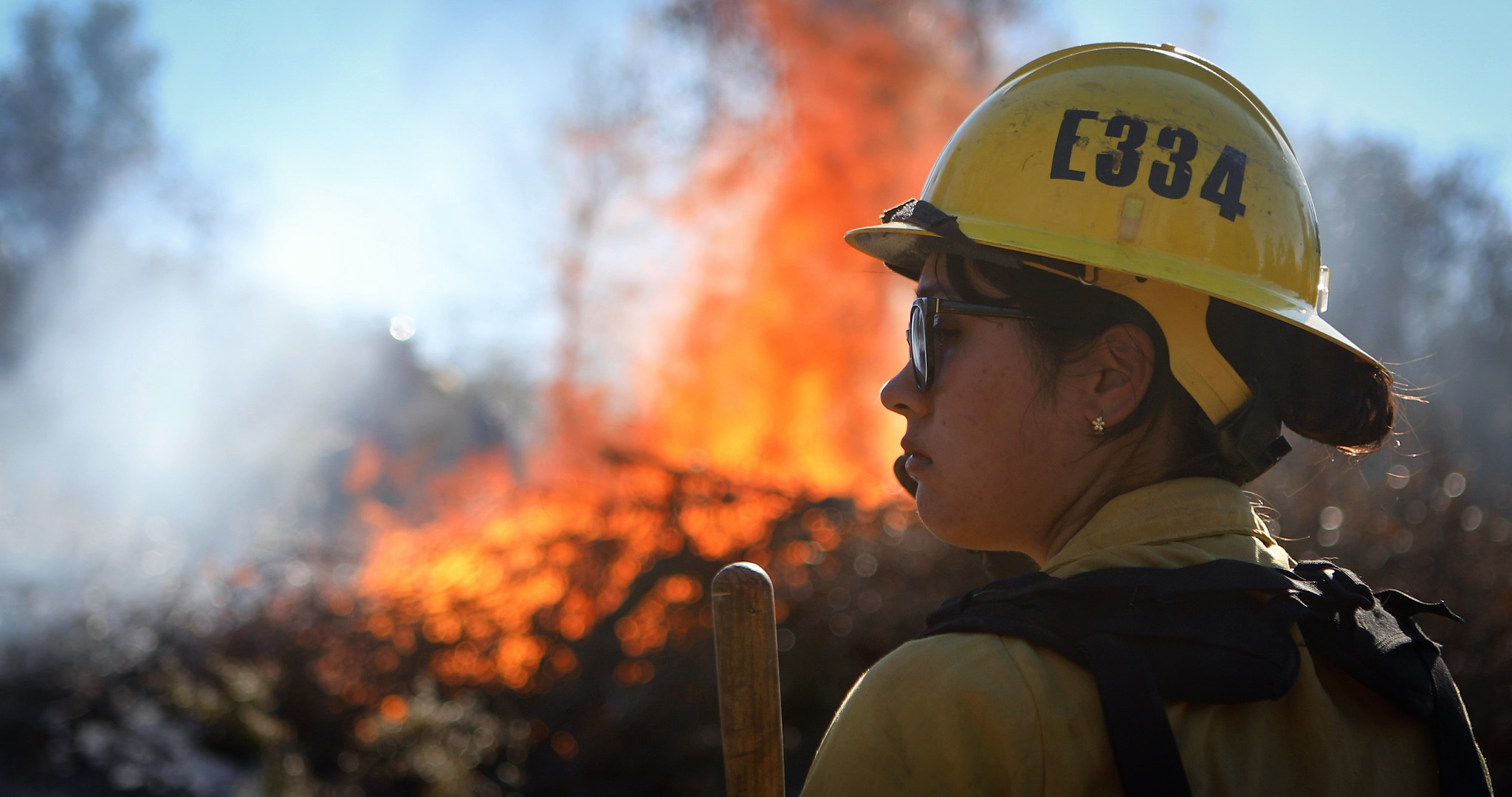 Firefighter in yellow hard hat with burning fire and brush in background.