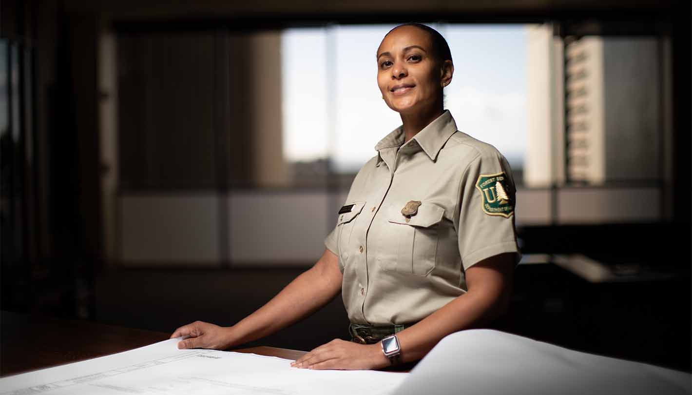 African American woman in tan Forest Service uniform rests hands on engineering drawings.