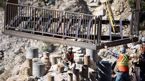 Forest Service contractors move a bridge onto pillars through a canyon using a crane.