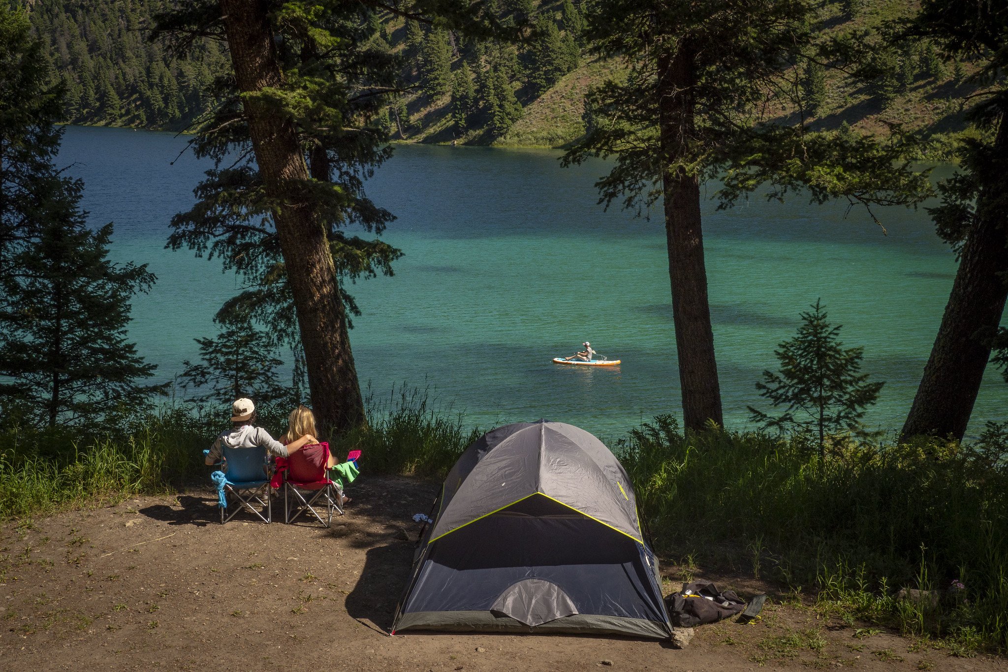 Two people sit by a tent at a forested lake.