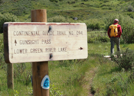 A picture of a person hiking the Continental Divide Trail, number 094. A sign on the trail shows the direction of Funsight Pass and the Lower Green River Lake.