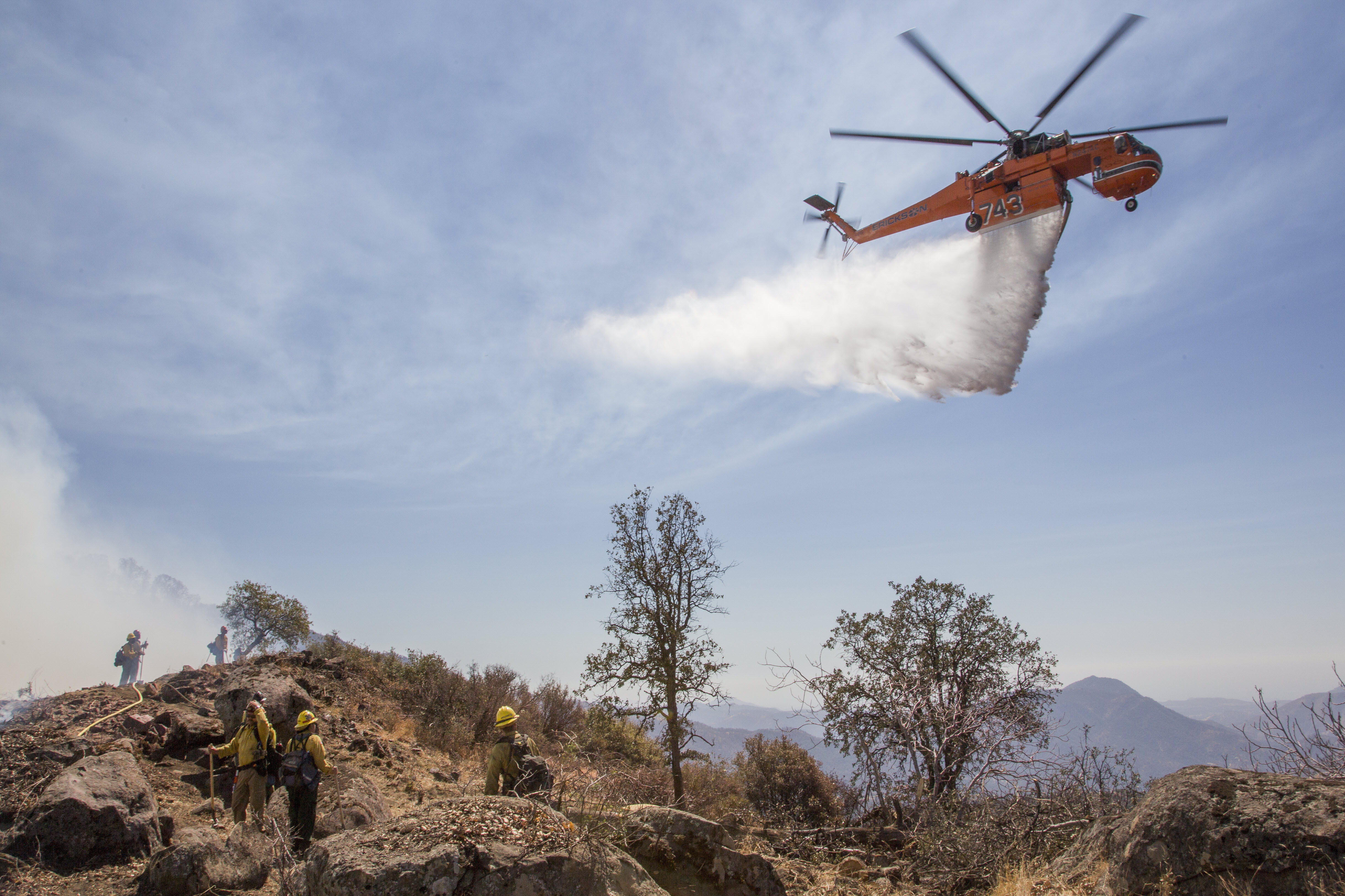A skycrane helicopter dropping water in the path of a forest fire, between the fire and a number of fire fighters working on top of a mountain.
