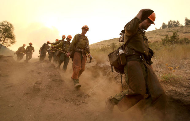 A line of wildland firefighters kicking up dust as they walk down a hillside fire break carrying equipment.