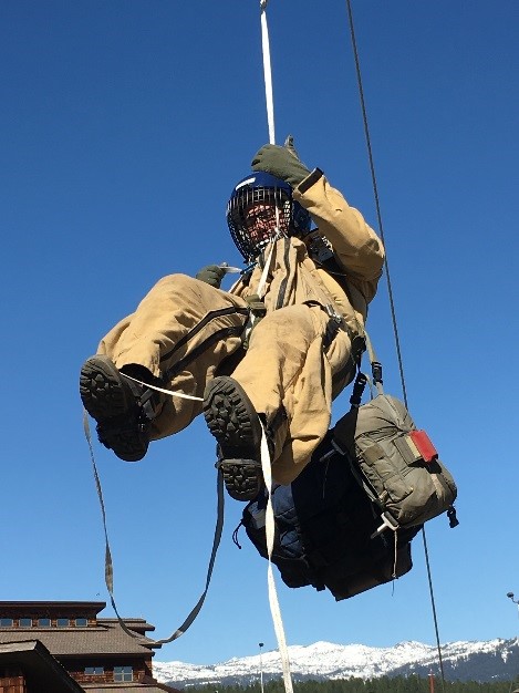 A smokejumper wearing full protective jump gear training to lower himself down from a simulated tree.