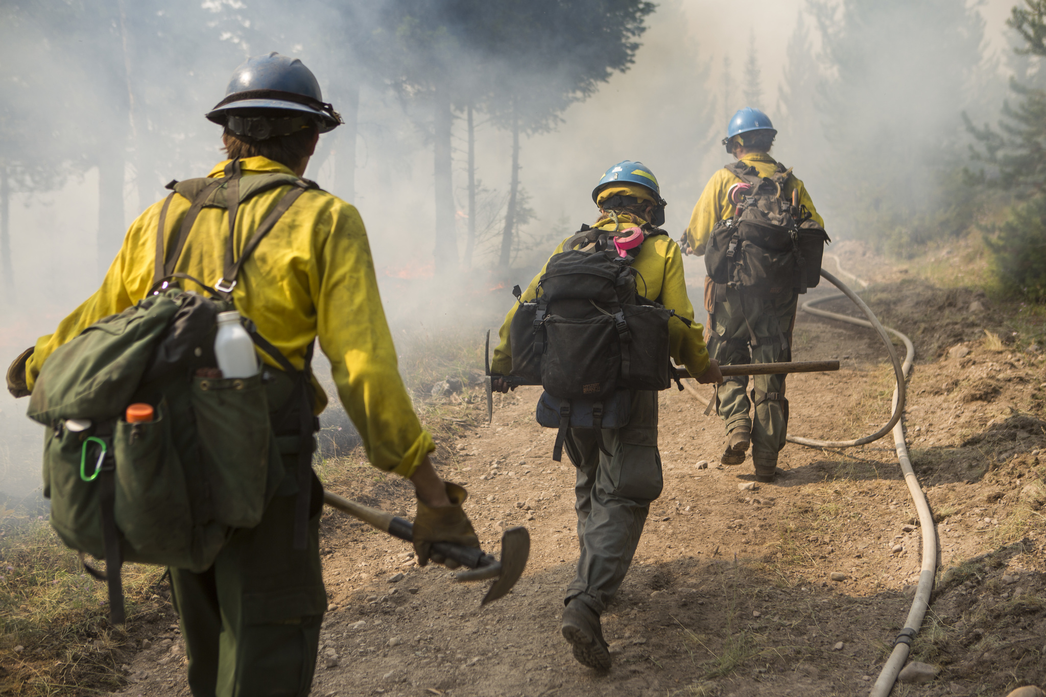 Three wildland firefighters walking along a cleared fire break, the first firefighter carring a fire hose.