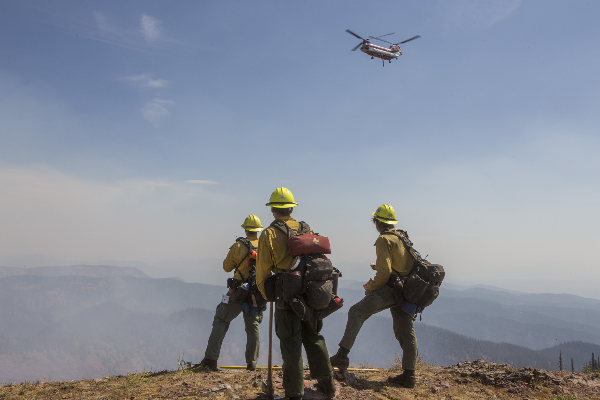 Three wildland firefighters standing on a high mountain ridge, watching a CH-47 Chinook helicopter water tanker on wildfire incident.