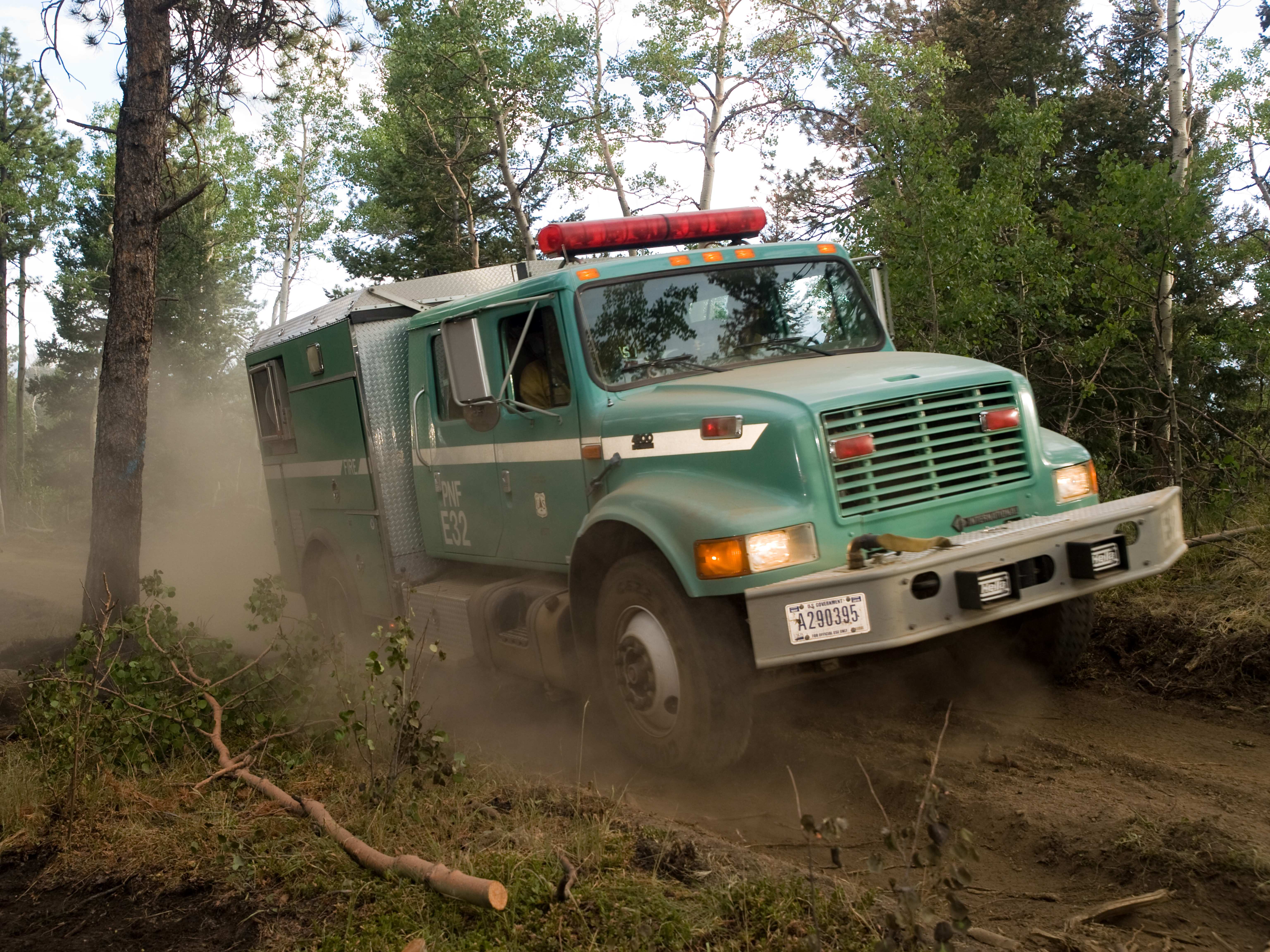 A green USDA Forest Service wildland fire truck moving down a dusty dirt road in a forest.