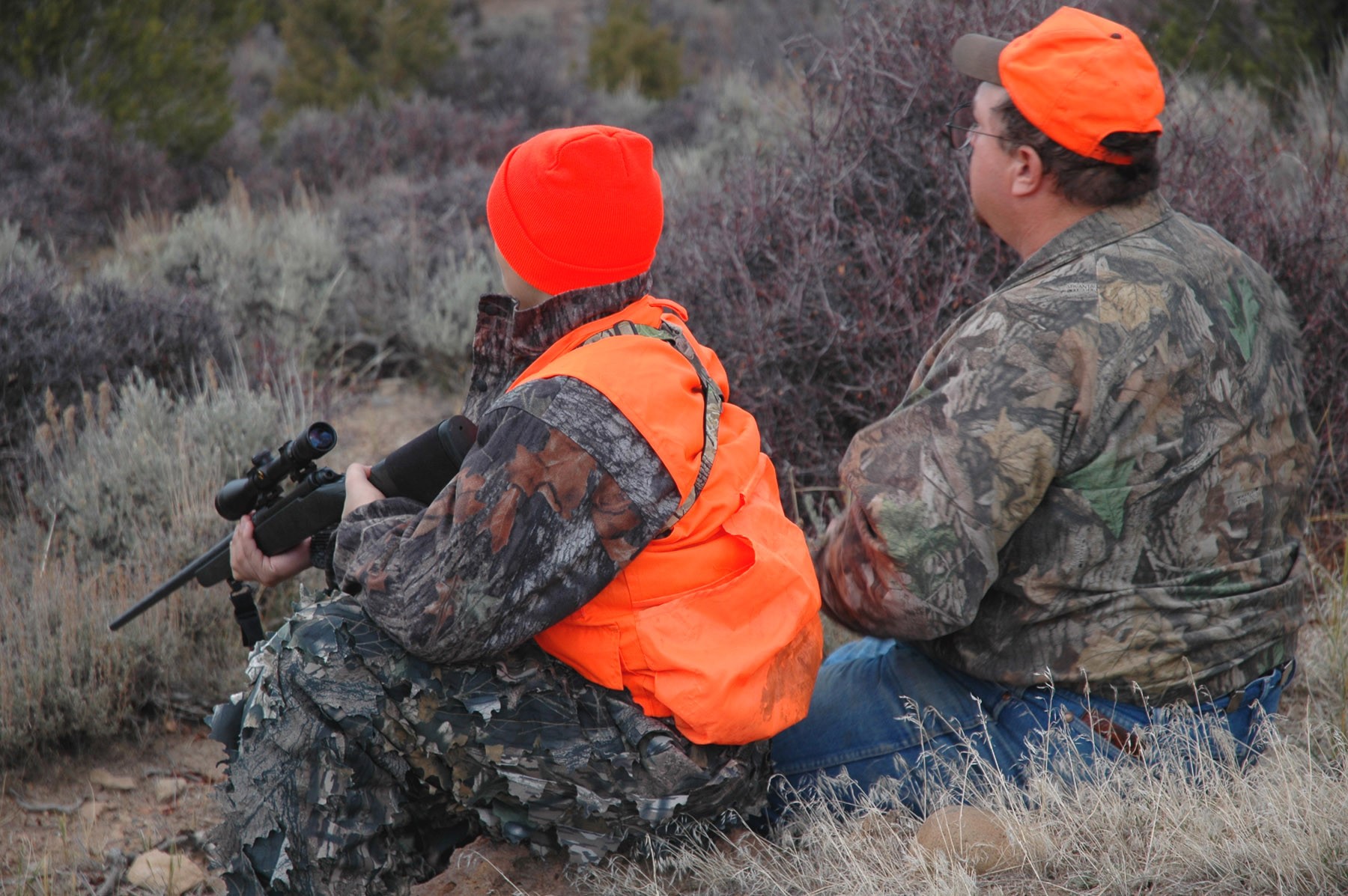 Two people sitting on the ground, holding hunting rifles, wearing camoflage clothing and orange high visibility hunting safety vests.