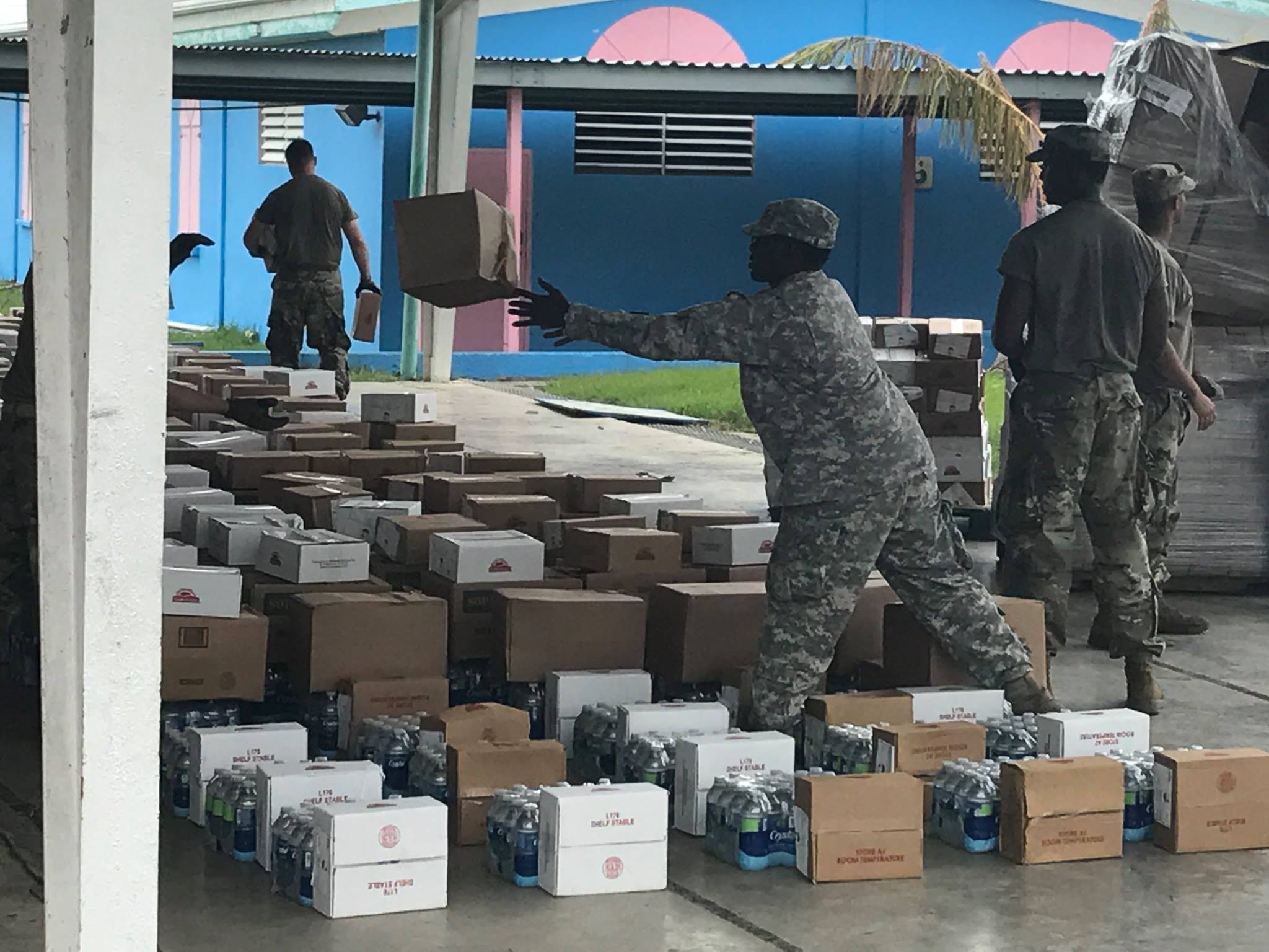 Soldiers in uniform arranging boxes of food and water under and awning. 
