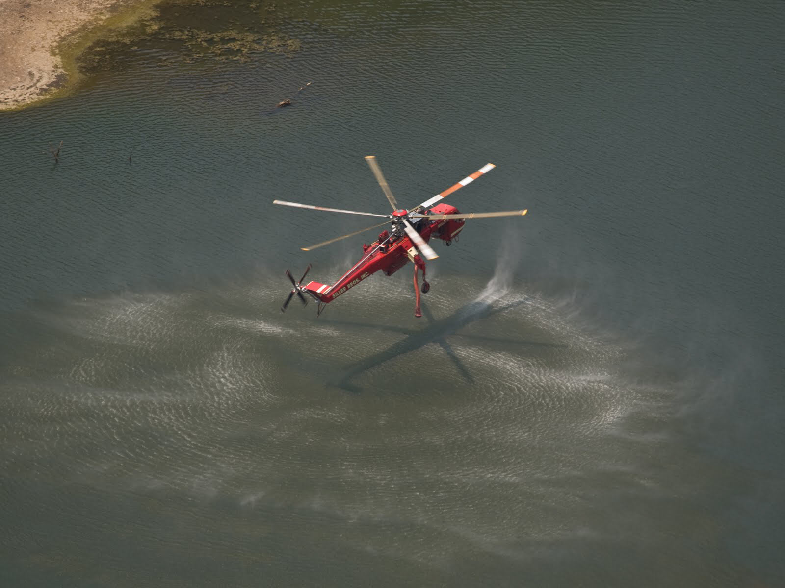 A sky crane helicopter hovering over a large body of water, preparing to dip a long hose to suck up water into a drop tank under the helicopter.
