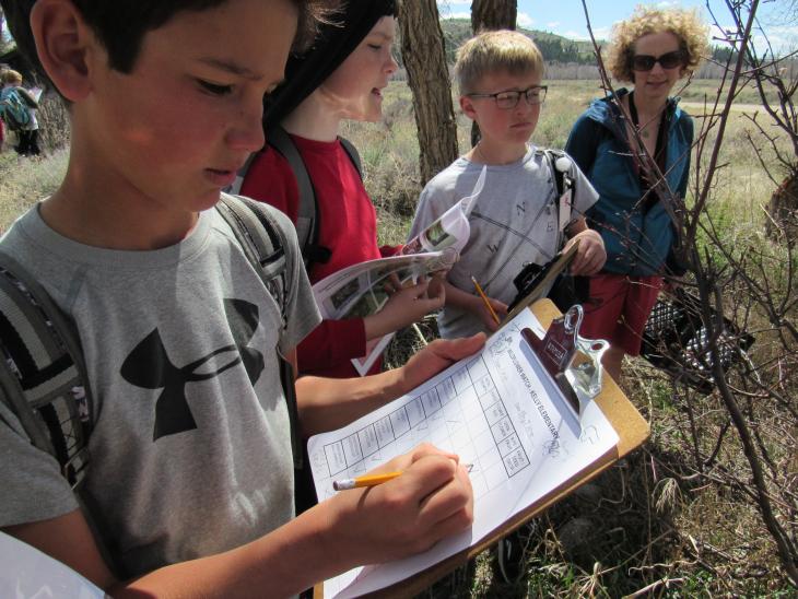 A row of three boys and one teacher take measurements of vegetation on a clipboard with mountains in the background.