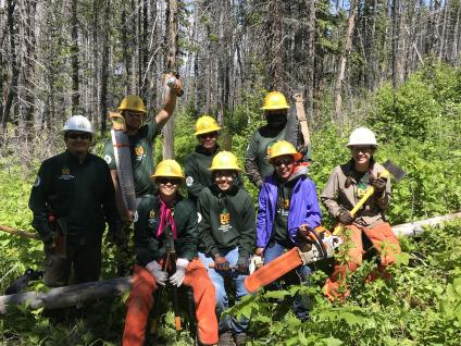 A group of smiling trail workers in a forest wearing hard hats and holding trail-building tools.