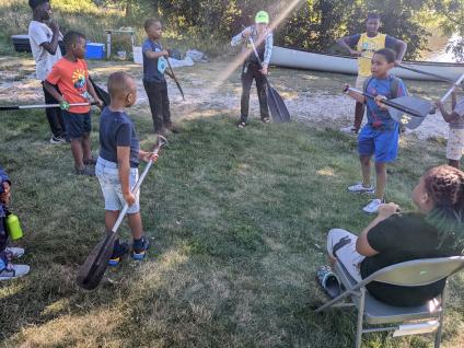 Young students stand in clearing holding canoe oars while an instructor demonstrates how to paddle.