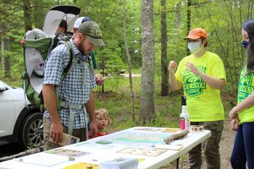 Two women wearing facemasks and yellow shirts assist a man wearing a hiking backpack while perusing maps with hiking information. The maps are set up on a table and all three are outside, in the forest