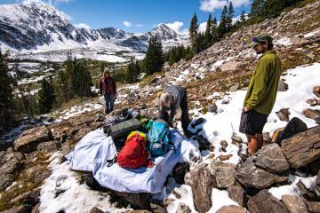 Three volunteers, two men and one women, stand on the side of a mountain, sparsely covered in snow, checkingt heir backpacks and equipment