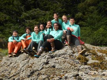 A group of nine young people sitting on a rock and smiling at the camera. They are all wearing pale blue/green long sleeved shirts.