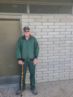 Older man, standing next to a doorway, cane on right hand. He is wearing a ballcap, sunglasses and a drak green windbreaker