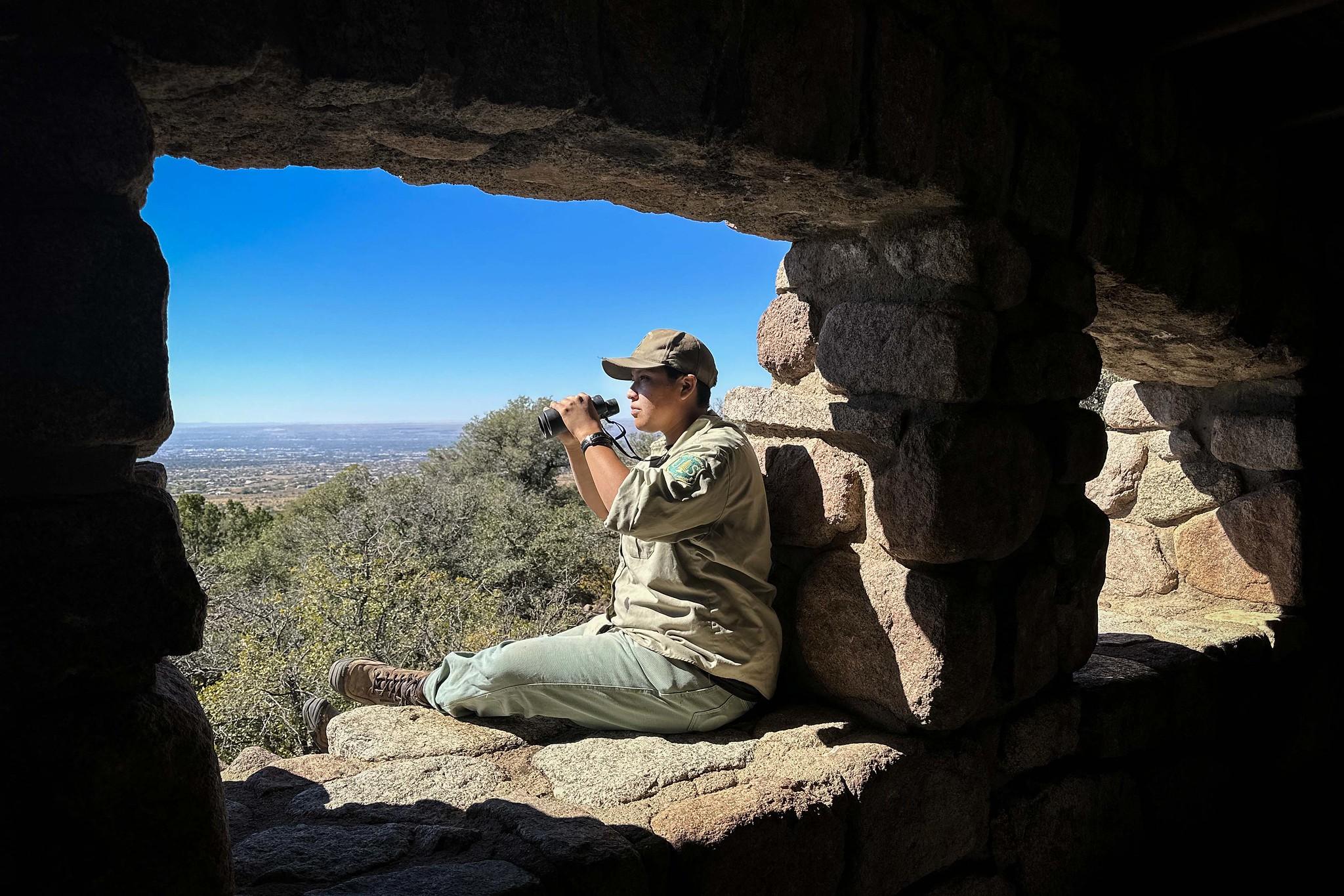 An employee sits in a window opening of a structure built of stone. They are using binoculars to look into the distance.