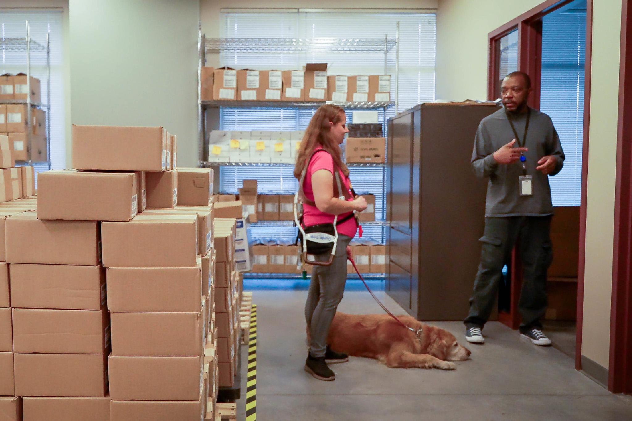 An employee with a service dog speaks with another employee in a warehouse filled with boxes.