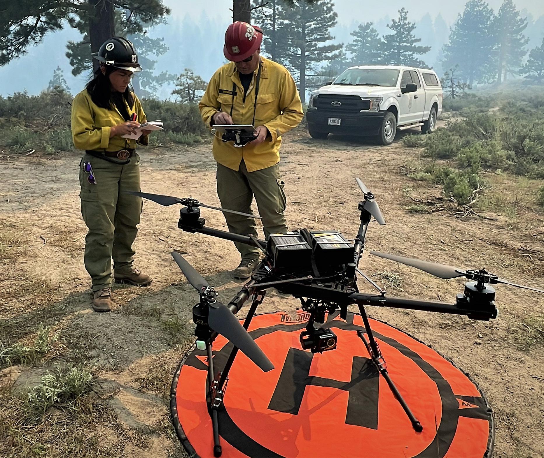 Two forest service employees stand next to a drone on an orange landing pad