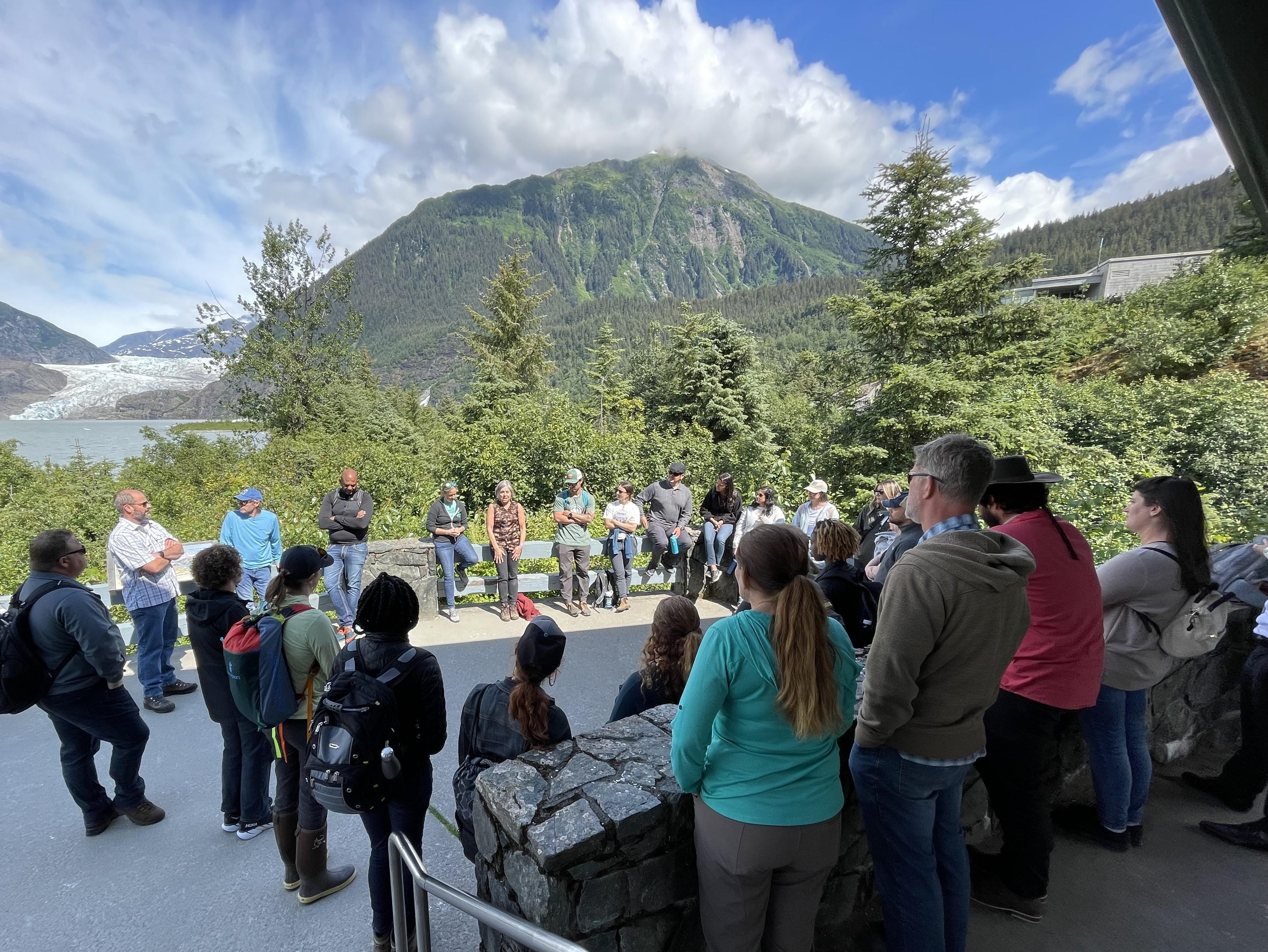 A group of people standing in a circle and talking in front of a background of trees and a glacier.