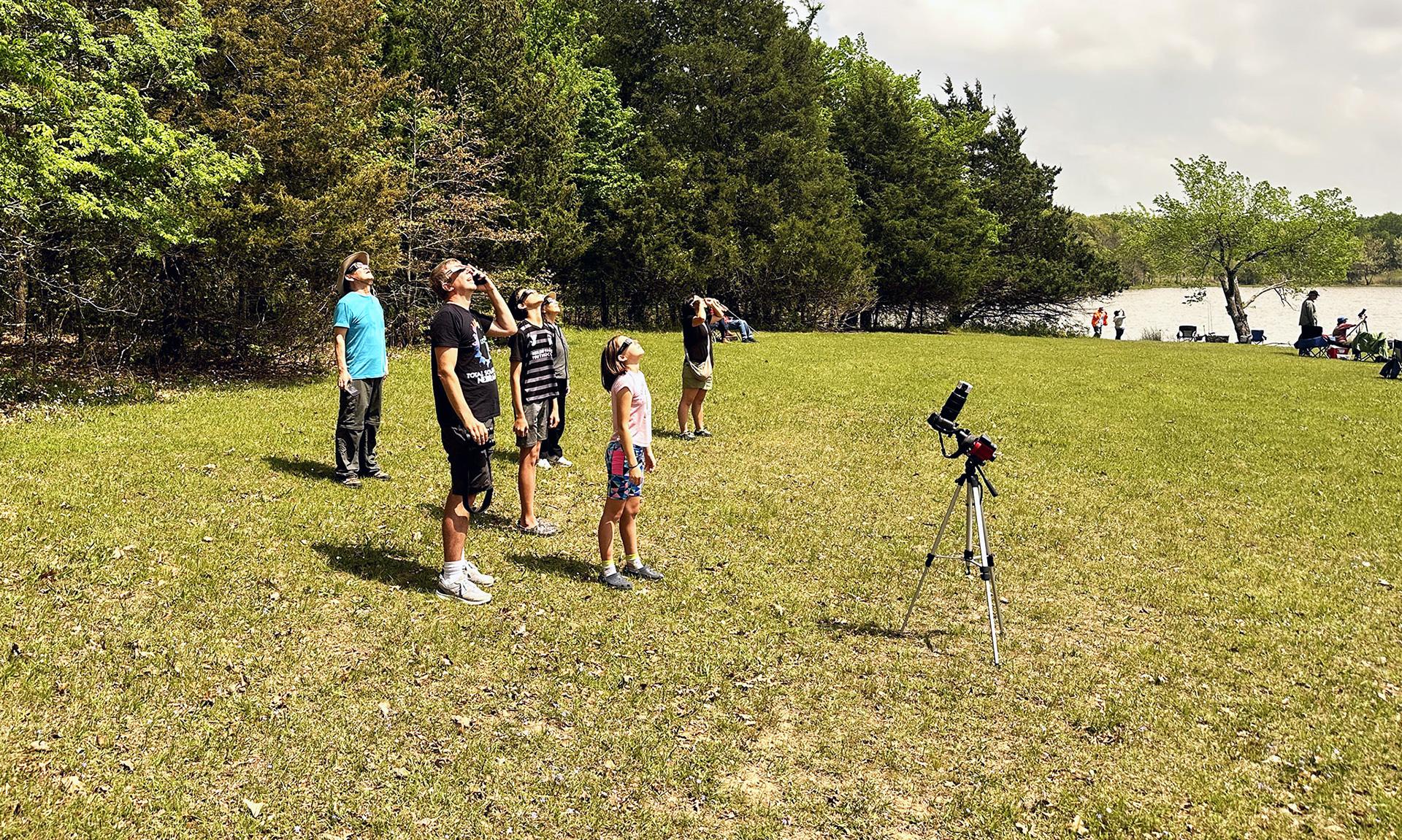 Children wearing Eclipse-viewing glasses while standing on a grassy plain, looking at the sky where the solar eclipse is happening.