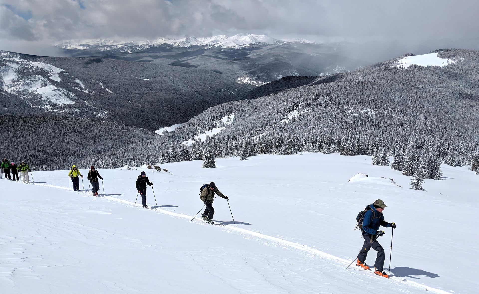 A line of cross-country skiiers trekking across a snow-covered plain with mountains in the background.