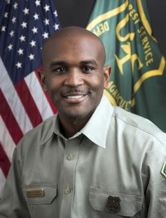 Portrait: John Crockett in Forest Service uniform in front of American & Forest Service flags.