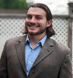 Profile photo of smiling man with shoulder length hair, sky blue shirt and tweet jacket