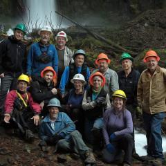 A group of volunteers wearing hard hats, posing for photo in front of a waterfall and forest debris.