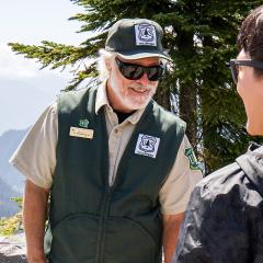 A volunteer wearing a USDA Forest Service uniform, hat, and sunglasses talking to another person.