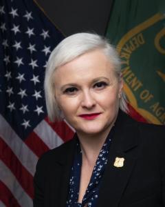 Portrait: Jaelith Hall-Rivera in suit jacket with small Forest Service lapel pin. In background, American and Forest Service flags.
