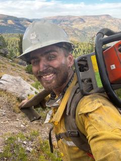 Simon Austin in nomex, hard hat, lugging equipment on a hillside. Face is slightly soot-covered. Mountains in background.