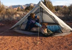 Camper in one-person tent still wrapped in sleeping bag. Sitting up and drinking coffee. Dog lying down beside them.