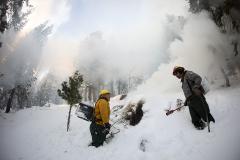 Two wildland fire fighters with propane torches standing next to a pile of limb debris burning under the snow.