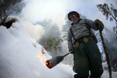 Firefighter, Adam Dummer walking with a lit propane torch next to a pile of limb debris burning under the snow.