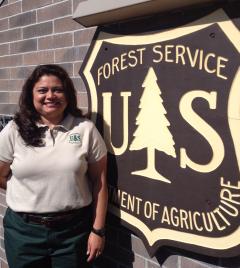 Betty Jewett, wearing Forest Service polo shirt and uniform pants, outside a brick building next to a large sign that is designed like the Forest Service insignia (badge).