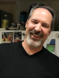 A man (shown from the chest up) smiling. He is wearing a black t-shirt, has a receding heairline and is sporting a salt and pepper goatee